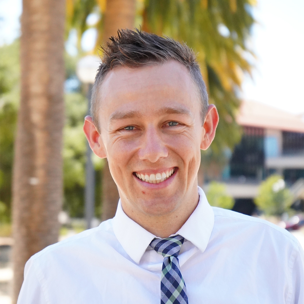 Headshot of John Hickey. He has short dark blonde hair, blues eyes, and light color skin. He is weating a white button down shirt with a blue and green plaid tie. He is standing outside on a sunny day with palm trees in the background.