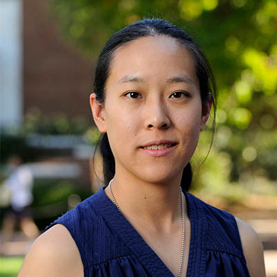 Headshot of Stephany Tzeng. She has long dark brown hair pulled back in a ponytail and dark brown almond shaped eyes. She is wearing a sleeveless v-neck blue blouse and silver bead necklace. She is standing outside in front of a red and white brick building and trees.