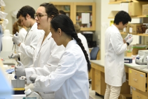 Wet laboratory setting. Three people in white lab coats are at a lab bench working on the right, while a student in the background works at a lab bench on the left.