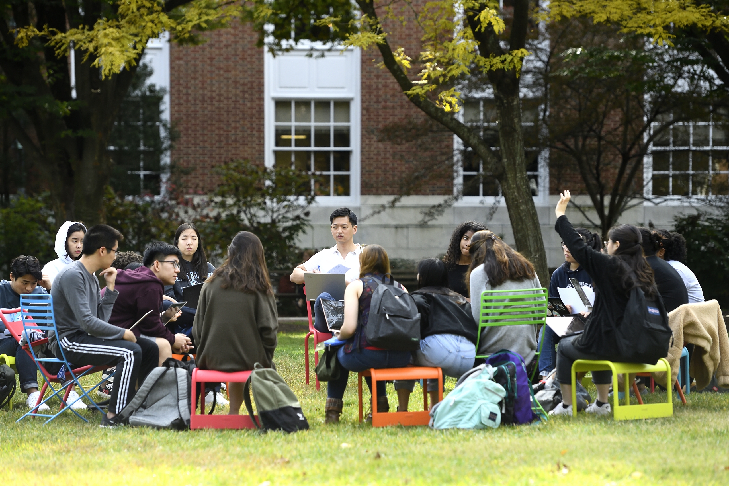 A large group of of about 12 students sitting in a circle on color metal chairs having a discussion outside on a college quad.